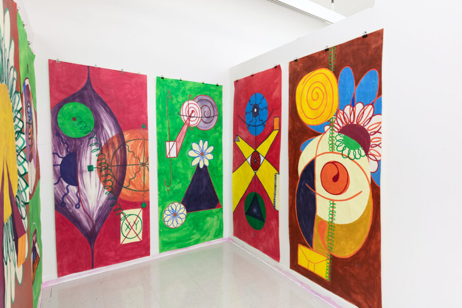 An installation view of six large, colorful paintings with geometric abstractions featuring images of eyes and flowers hanging in a white-walled studio.