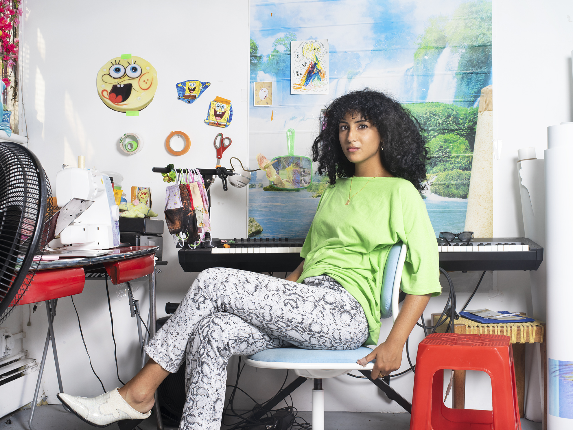 A photograph of Farah Al-Qasimi in her studio. Farah is seated on a chair in the foreground looking at the camera. There is a piano keyboard behind her and other ephemera hanging on the wall behind her.