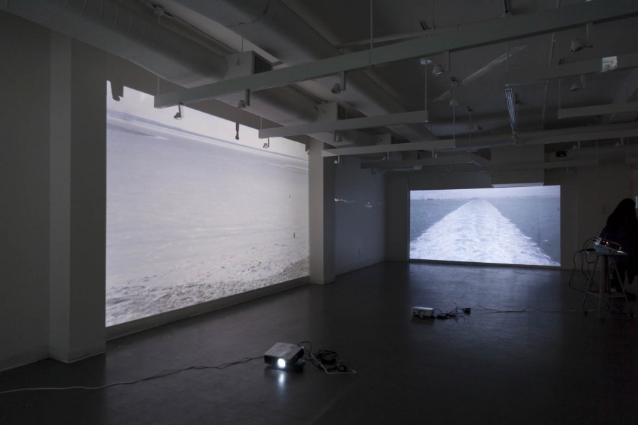 Installation view of two image projectors that are projecting different images from "Europe's Ice core Sampling". On the left is a clear ocean view, and in the background is a view of a boat trail on the water.