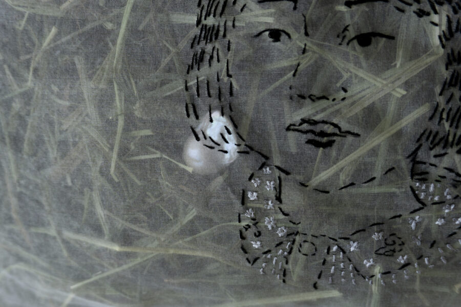 A pillow constructed of translucent white organza and stuffed with green straw. I portrait is embroidered on top of the pillow in black thread. This image shows a close-up of the embroidery stitching.