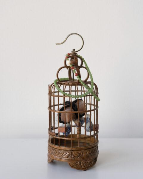 A small metal and bamboo cage with a hook on top. A string of jade beads wraps around the top and inside is an arrangement of human hair and other materials.