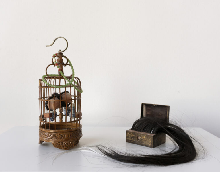 Two small sculptures on a white pedestal. The left sculpture takes the form of a small cage with a jade necklace draped over it. The other is a small, ornate metal box with a sweep of black and silver human hair emerging from within.