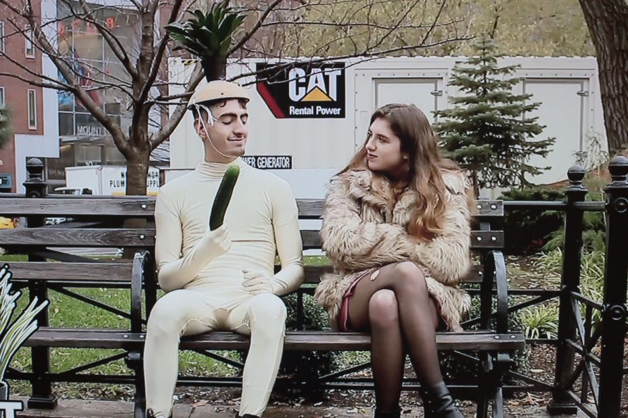 Video still of a man in a white bodysuit holding a cucumber wearing a funny hat sitting on a park bench with a woman in a fur coat.