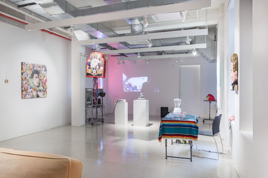 Installation shot of a gallery space with a table with a striped blanket on it and various sculptures in the space with pink lighting in the background.