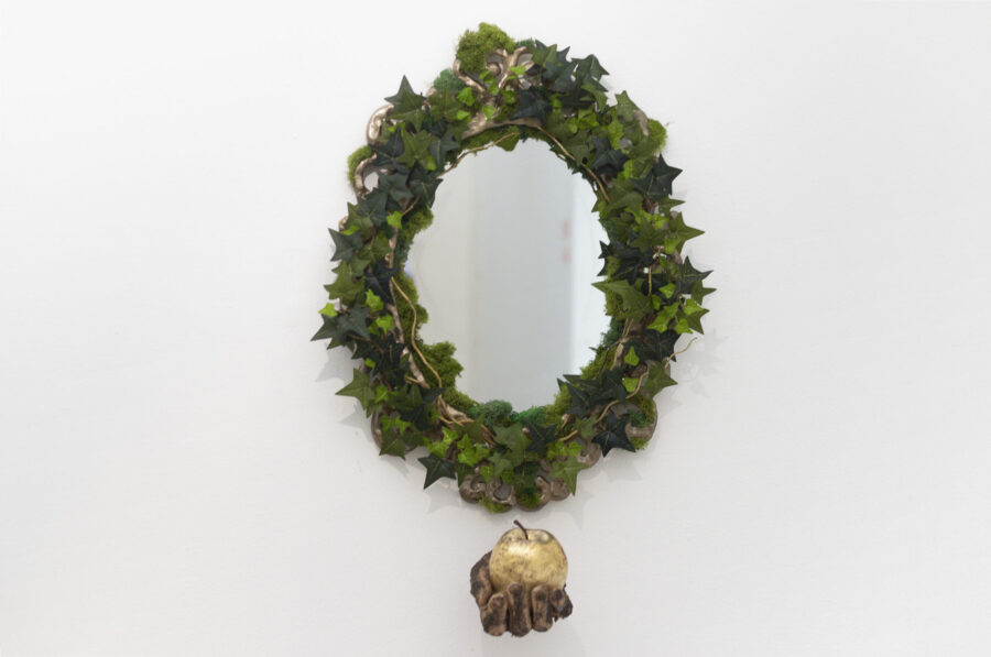 A round ornate gold mirror covered in artificial ivy and moss, below the mirror is a hand facing the viewer holding a golden apple. The hand is covered in dirt.