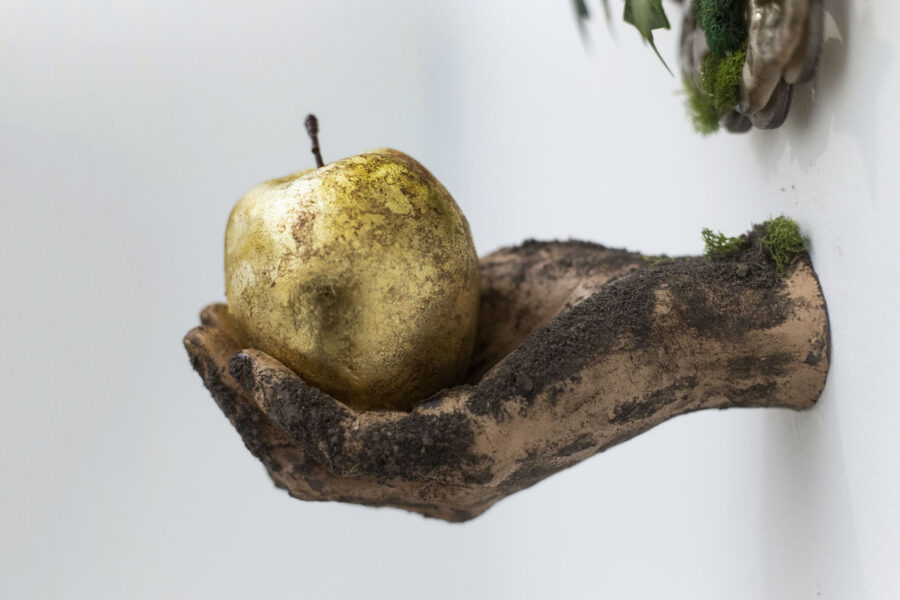 Detail shot. A round ornate gold mirror covered in artificial ivy and moss, below the mirror is a hand facing the viewer holding a golden apple. The hand is covered in dirt.