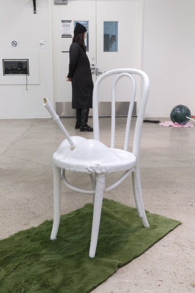 White chair with two rounded shapes and a whistle between the shapes, and a person dressed in black standing in the background.