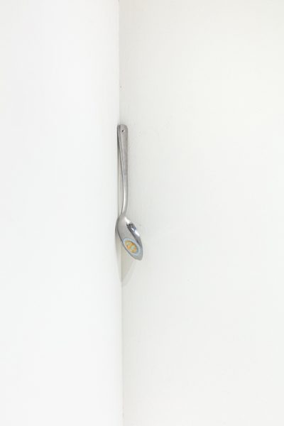 A sculpture by Hanjun Zhang. A silver spoon with a coin attached mounted on a white wall.