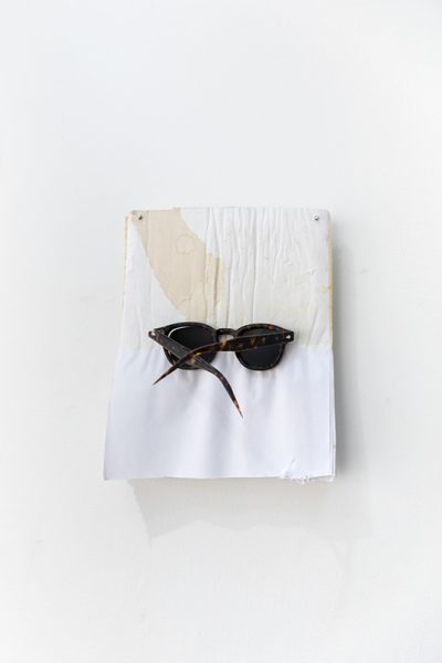 A mixed media artwork by Hanjun Zhang. Sunglasses attached to fabric mounted on a white wall.
