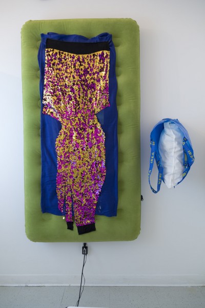 A green inflatable mattress on the wall with violet and golden sparkle material on blue fabric and a bag with a pillow beside it.