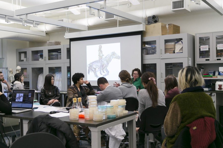 A class overview with students sitting on desks, a table with working materials in different rounded shaped recipients, many cabinets with glass doors and objects in them, and a projector surface with an image projected on it