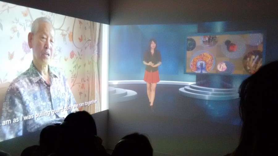Installation view of images projected, one with an older man and a caption on the lower part of the image, and another with a woman presenting news. In the room are a couple of people watching the projections