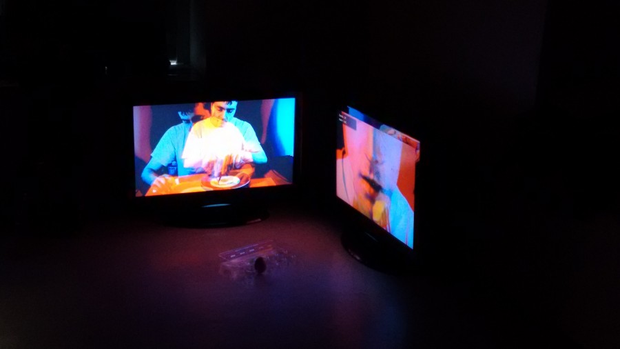 Installation view of two TVs displaying stereoscopic images of a man on a table, and another image with a close-up to his mouth