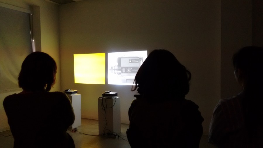 Installation view two video projectors on stands, one is projecting a yellow image on the wall and the other one, a picture with a car, while in the room are three people looking at the photos on the wall