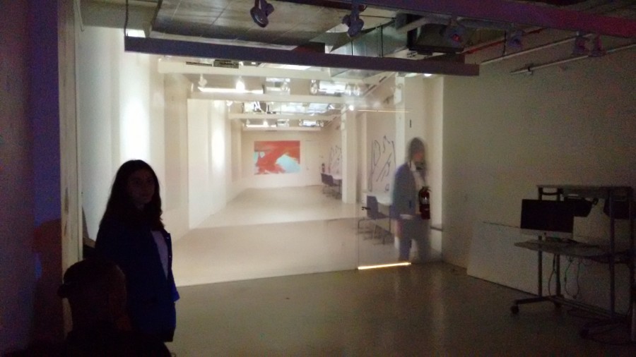 Installation view of a video projector and an iMac on a desk near the right wall, and an image projected on the wall with a room and a person on the right side. In the installation room is a person facing the camera in the lower-left corner