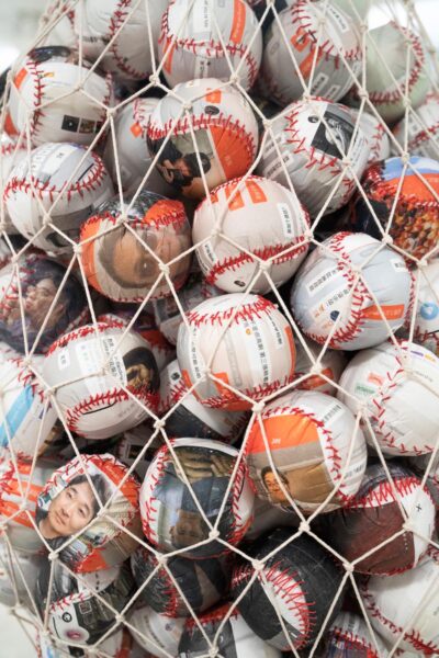 A detail of An's artwork. A net suspended from the ceiling holding a bunch of baseballs. Each ball is made up of printed material that has different faces, text, or screenshots printed on them.