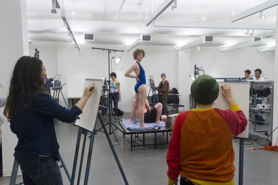 A drawing class with models. In the center of the room are two models on the platform. One is sitting. The other is standing. In the background, around the room, students are standing at their easel and drawing the models.