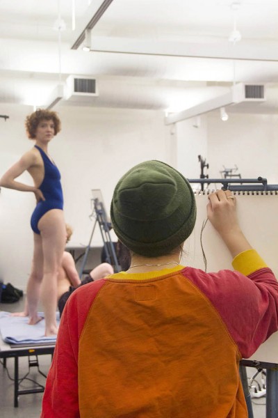 A student in the midst of drawing a still life from a model. In the foreground of the image a student is faced away from the camera and is drawing on their easel. In the background, to the left, are two models on a platform. On model is standing and wearing a blue swimsuit. The other model is sitting and wearing dark shorts.