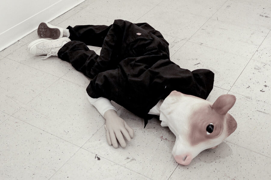 A human figure wearing white sneakers and black clothes with a plastic cow head lies on a linoleum-tiled floor.