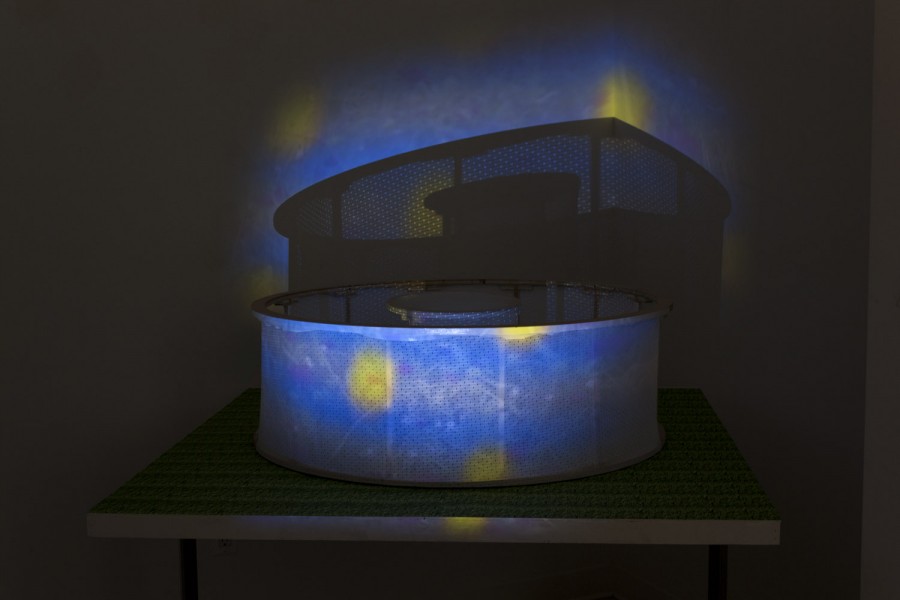 Ceramic sculpture installed in a rounded recipient with a fine mesh, in a dark room, an image is projected on the sculpture with blue background and yellow dots