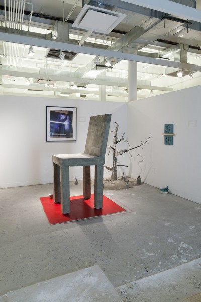 Installation view of a giant chair on a red squire and different objects.