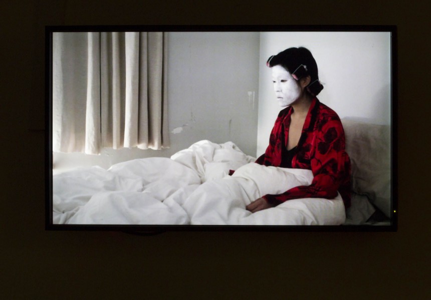 A person sitting in bed with a white pillow over the feet, dressed in a red shirt with black spots, has the face painted in white. In the background are a window curtain on the left side and a small amount of light coming from the left side.