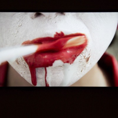 Screen capture from "The Beach elorette Party" with a face painted in white, red lips and a toothbrush cleaning the teethes and red a red-colored fluid coming out from the left side of the lips.