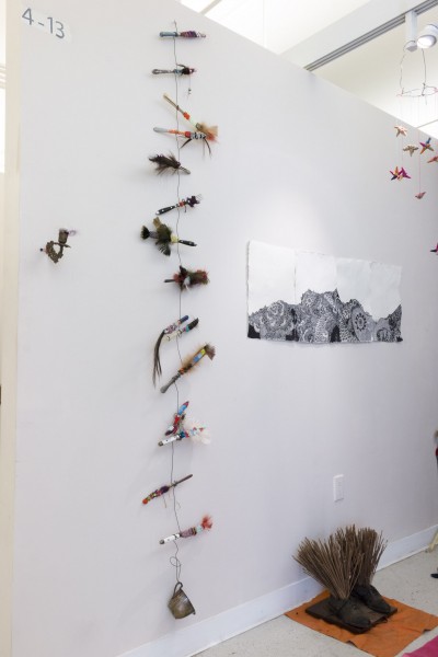 Miscellaneous objects are hanging on the wall and a print with black mandala patterns on white background, and a pair of shoes on the floor with many thin wood sticks in them.