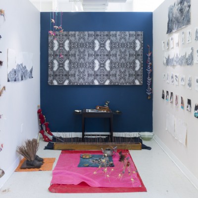 Installation view of structures made of thin wood sticks, a shattered plate, a teapot, many miscellaneous objects, and vibrant colored fabrics, and other objects hanging on the wall.