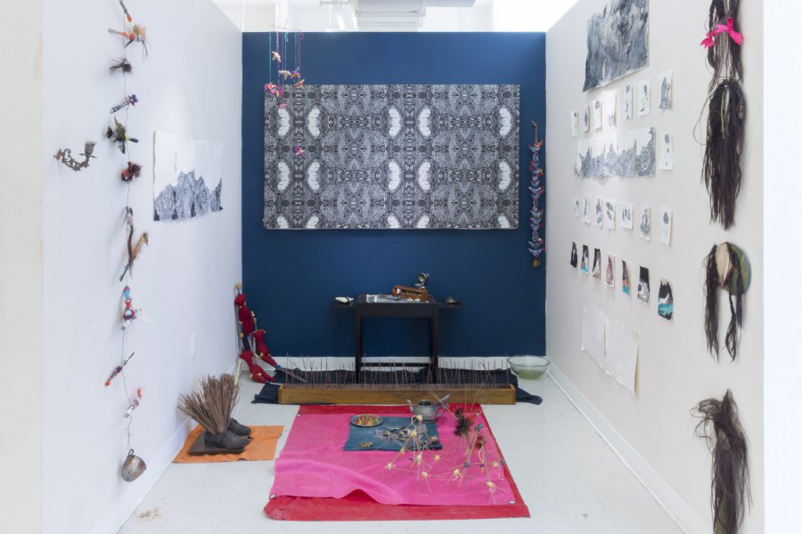 Installation view of structures made of thin wood sticks, a shattered plate, a teapot, many miscellaneous objects, and vibrant colored fabrics, and other objects hanging on the wall.