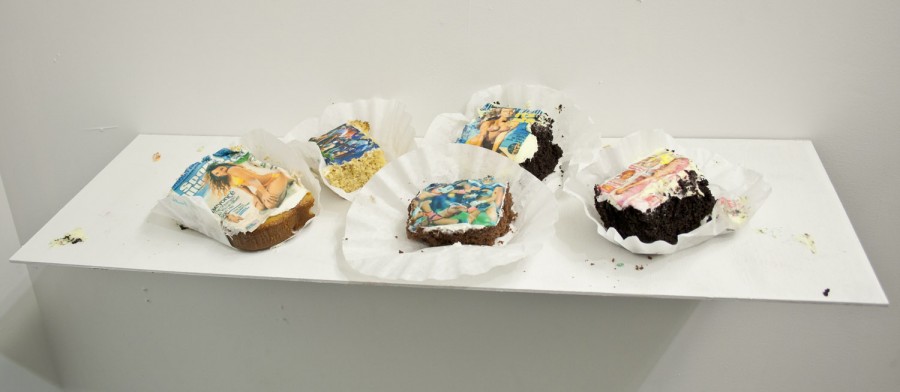 Five cake pieces with images of a magazine cover with a woman in a bikini, a football game scene, another magazine cover, etc., cakes are packed in individual cupcake paper sheets on a white shelf.