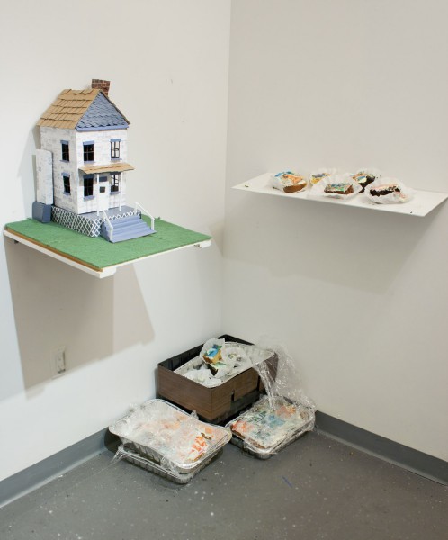 Installation view of a house with brown rooftop and blue windows, and blue stairs, on a green matt installed on a shelf, another shelf with rounded organic shapes inside cupcake cooking paper, a basket with different organic material, and four trays with paints and other materials.
