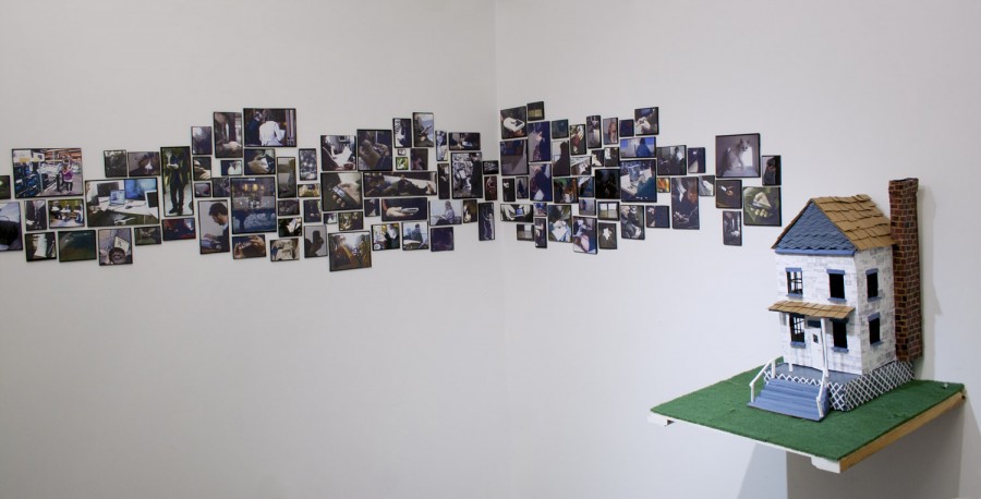 Installation view of a masonry grid of photos and a small house sculpture placed on a shelf with green fabric on it