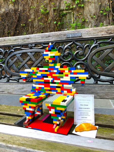 Man made of Legos sitting on park bench next to a croissant.