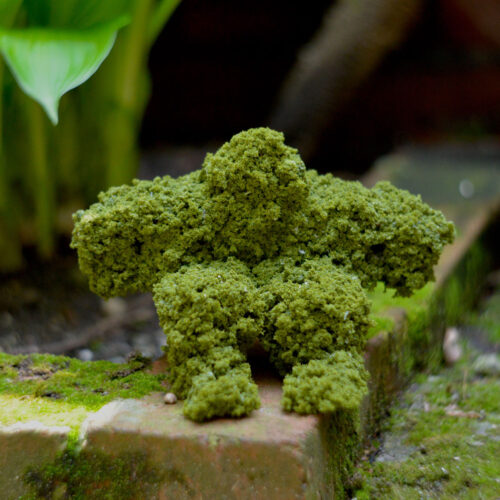 Small man made of moss sitting on ground next to a green plant.