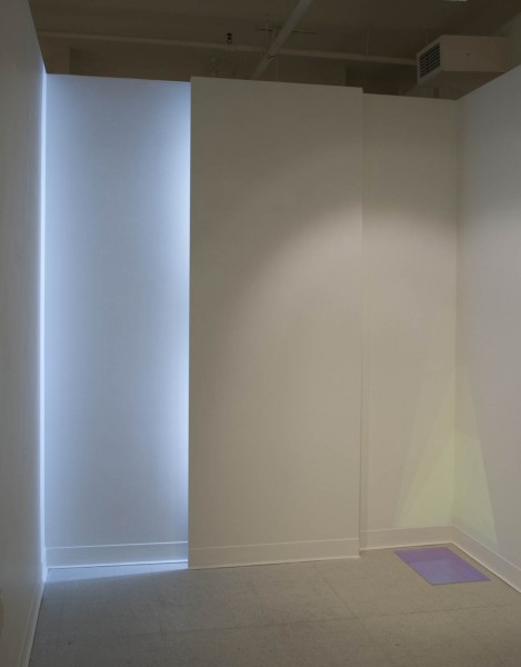 Installation view of a wooden structure with a bright light behind a wood illuminating one side of the wall and an acrylic sheet placed on the ground near a wall corner reflecting colors on the corner.