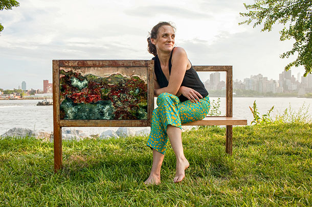 Portrait of Jenifer Wightman sit down on a park bench. Next to the artist there is a painting. The background incudes the skyline of a city.