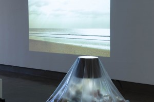 A house lamp is placed on a rectangular shape on the floor, surrounded by trash and covered with transparent plastic vellum. In the back is a projection of a beach and ocean with big heavy clouds in the sky.