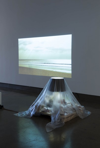 A house lamp is placed on a rectangular shape on the floor, surrounded by trash and covered with transparent plastic vellum. In the back is a projection of a beach and ocean with big heavy clouds in the sky.