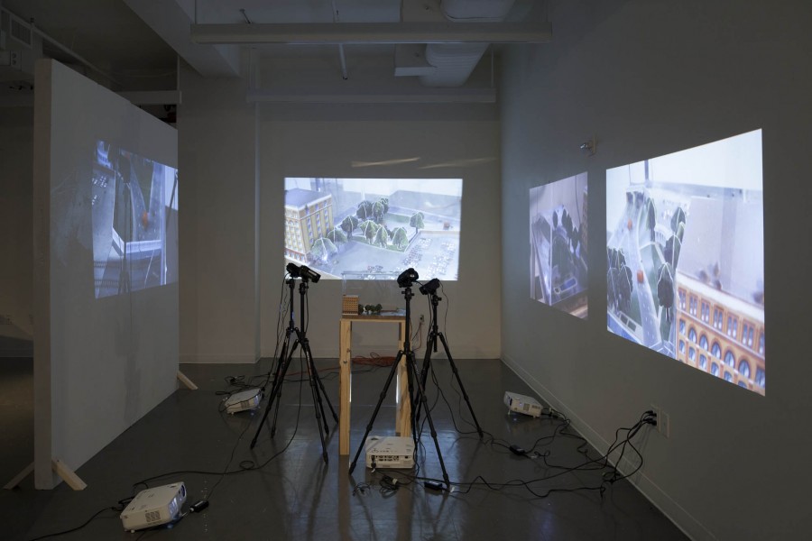 Installation view of a model in an aquarium with a tall yellow building, green trees, and grass filmed by four cameras and two images projected on the left, in the center, and right walls