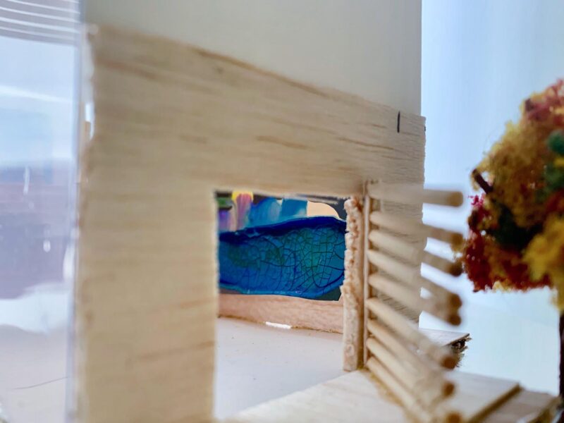 Cardboard and wood model of a home with a swinging wooden door and red tree outside.