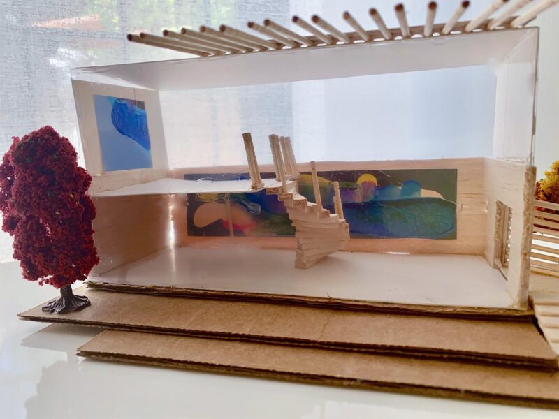 Cardboard and wood model of a home with a swinging wooden door and red tree outside and a spiral staircase inside with a blue model painting. 