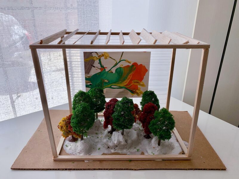 A wooden model home with a miniature flowery painting with nine red and green trees placed in sand in the interior.