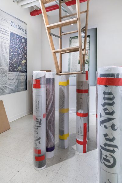 Installation view of artwork by Jisoo Na. Print of a newspaper article, rolls of printed images with yellow and red tape and an upside down ladder hanging from the ceiling.