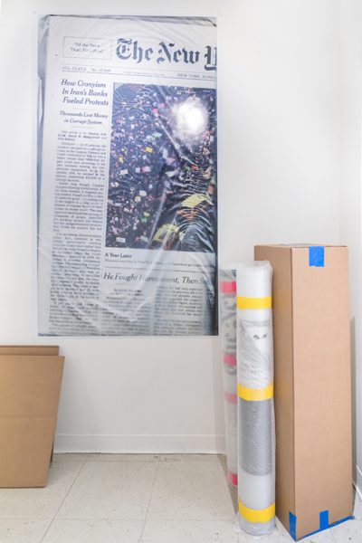 Installation view of artwork by Jisoo Na. Print of a newspaper article, rolls of printed images and a box with blue tape.