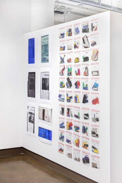 Installation view of artwork by Jisoo Na. Grid of plastic resealable bags with various colored geometric shaped items inside along with newspaper clippings mounted on a wall..