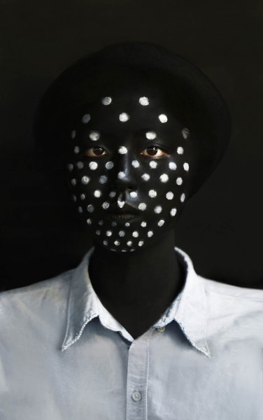 Headshot portrait of a person with a black hat and with skin painted in black and with silver dots on the face