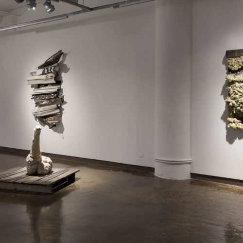 Installation view of an organically shaped sculpture in the middle of the room on a wooden stand, two other sculptures hung on the wall made with angled shapes, and another one on the far right with rounded shapes made of a white organic material