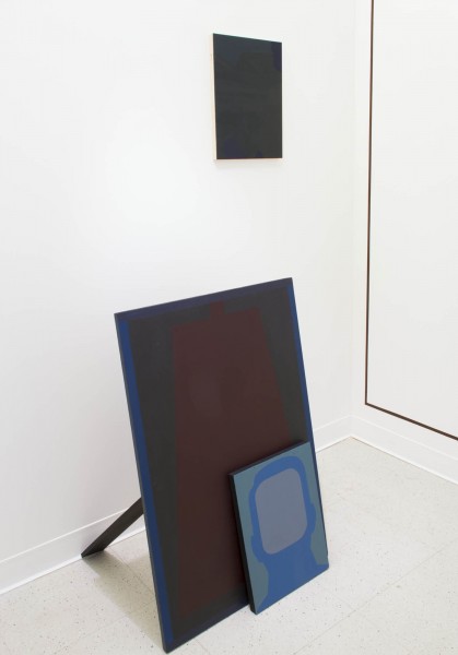 There is a side view of three paintings. Two paintings are placed in front of the other, where a smaller painting with a blue silhouette portrait is put in front of a bigger dark red silhouette portrait. On the wall, in the background, is another painting.