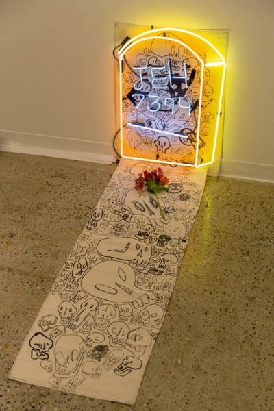 Installation of a tombstone rock made of a yellow neon as an outline, and the text JEH 7934 is written with white neon. At the base of the neon is a long piece of paper with skulls drawn on it and a red flower